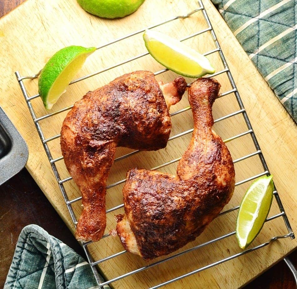 Top down view of 2 crispy chicken leg quarters on top of wire rack with lime wedges on top of wooden board.