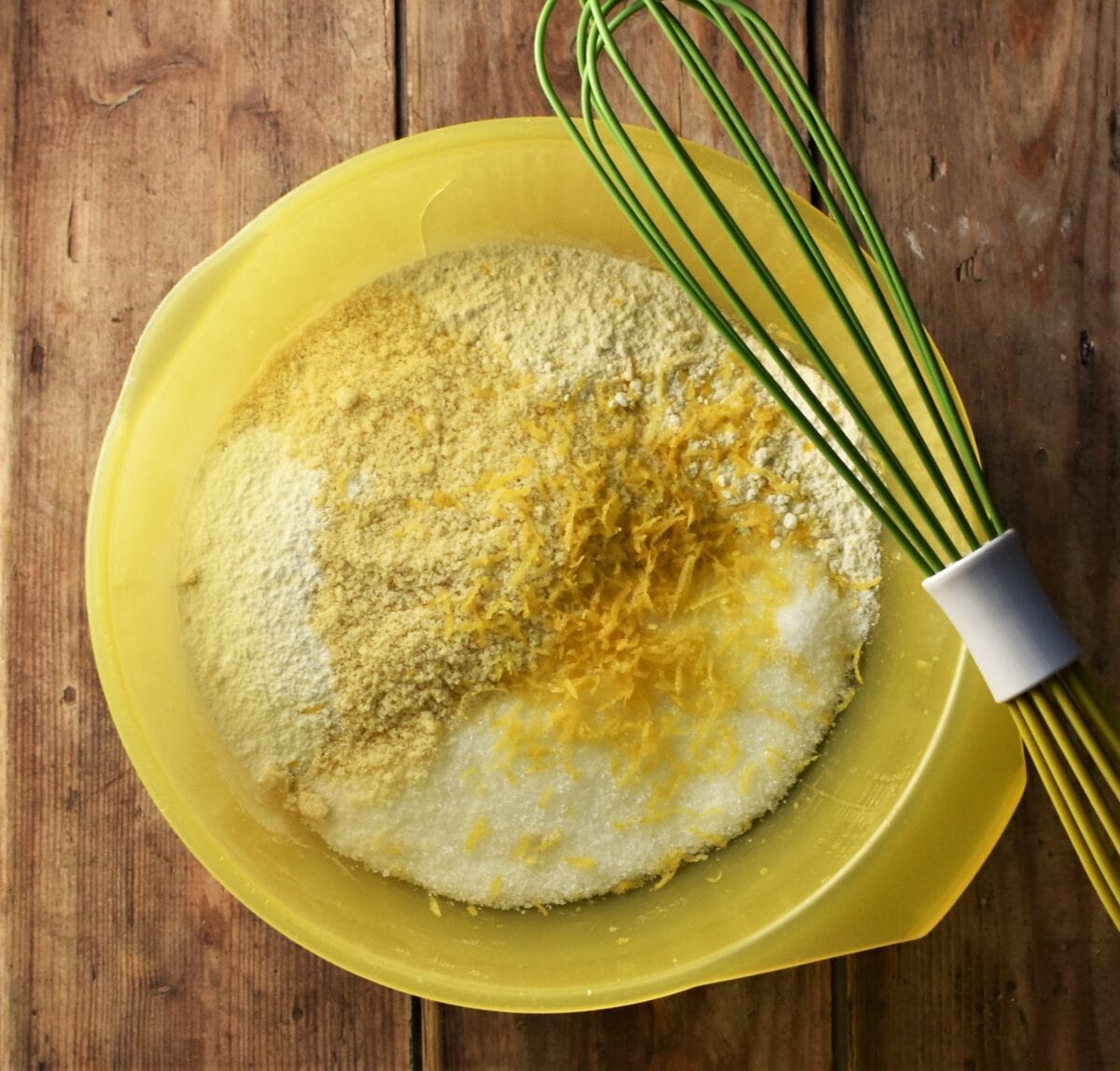Flour mixture and lemon zest in large yellow bowl with green whisk on top.