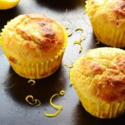 Close-up view of lemon muffins in yellow paper cases with lemon zest on top of dark surface.