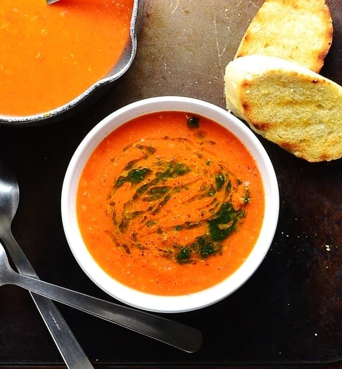 Top down view of tomato soup in white bowl with spoons, toasted bread and soup in saucepan in background.