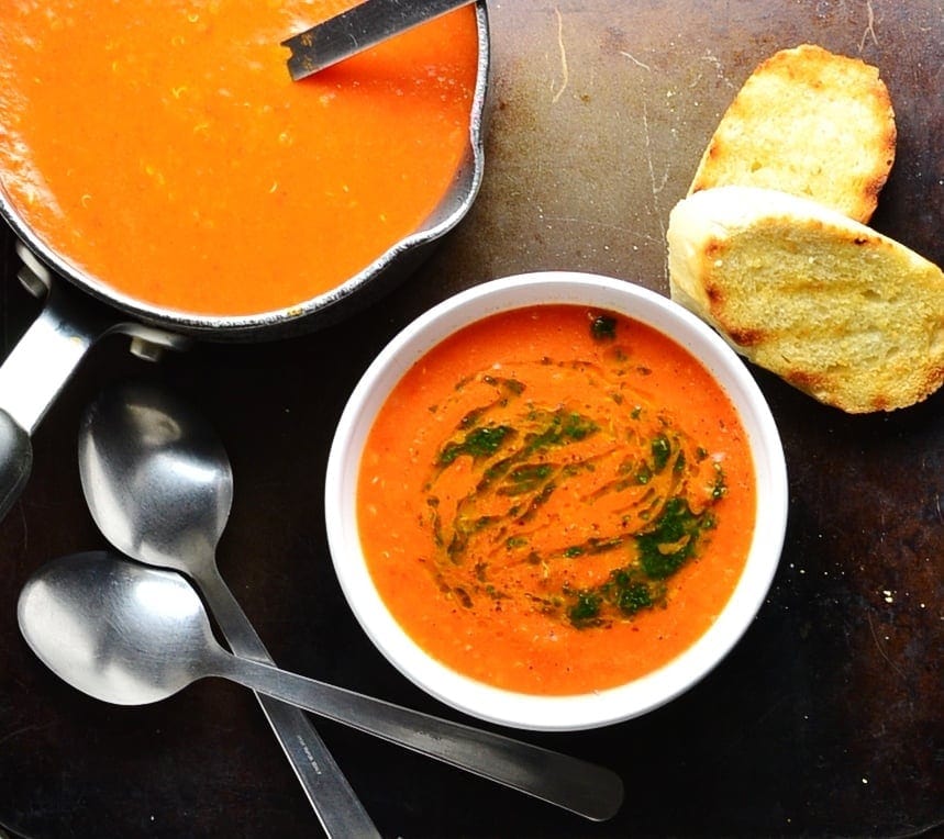 Top down view of tomato soup with basil drizzle in white bowl and black saucepan in top left corner, toasted baguette slices and spoons on black table.