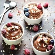 Chocolate banana pudding in 3 white dishes with spoons, fruit and chocolate shavings on dark table.