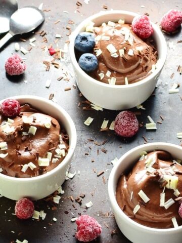 Chocolate mousse in 3 white dishes with spoons, fruit and chocolate shavings on dark table.