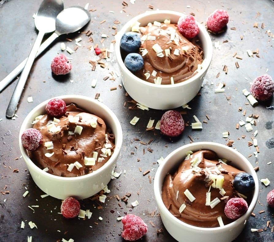 Chocolate mousse in 3 white dishes with spoons, fruit and chocolate shavings on dark table.