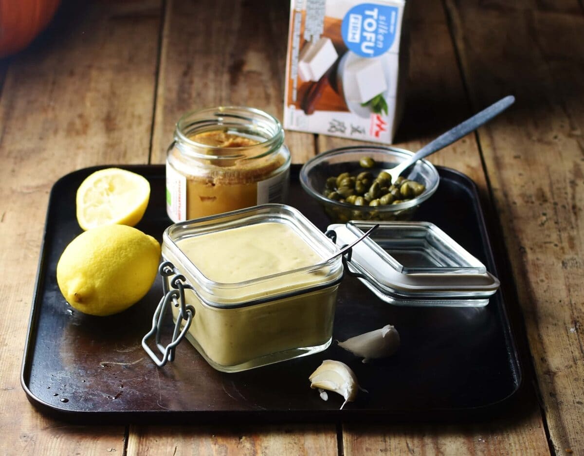 Dressing in square open jar with spoon, lemon, miso paste in jar, capers in small dish with spoon and silken tofu package in background.