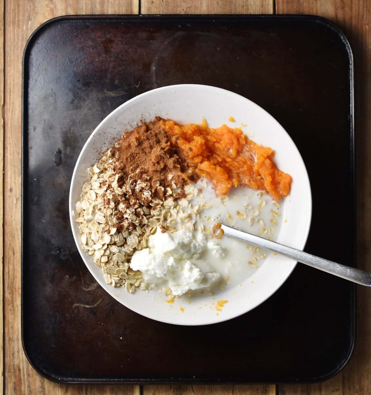 Top down view of oats, milk, spice, yogurt and mashed sweet potato in white bowl with spoon.