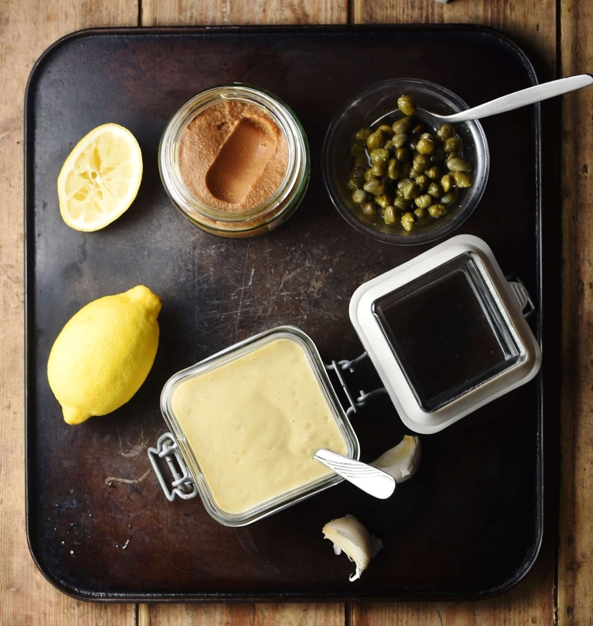 Top down view of caesar dressing in square jar with spoon, lemon , miso paste in jar and capers in small dish with spoon.