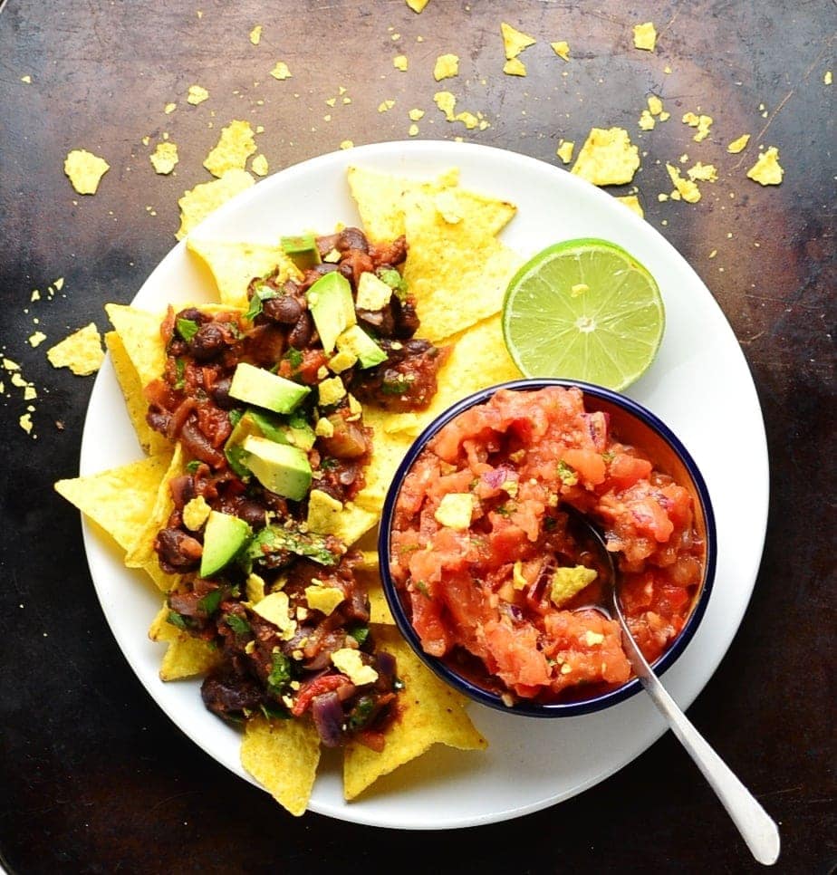 Mexican black bean stew on bed of nachos with salsa in blue dish with spoon and half lime on white plate.