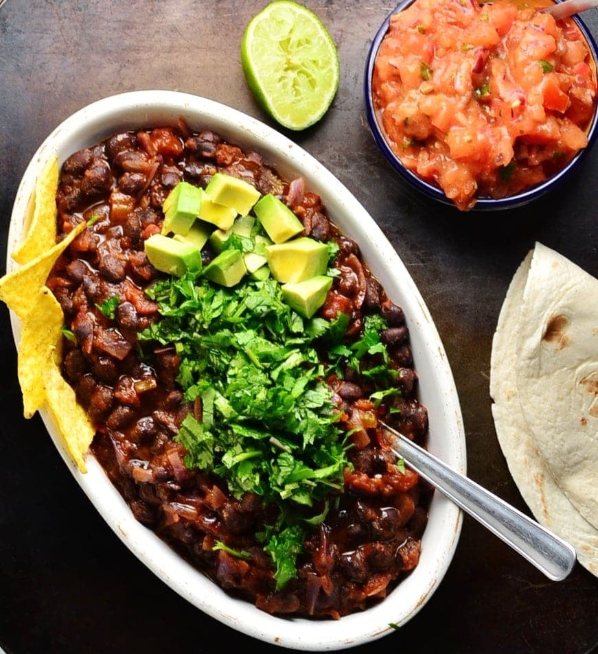 Black bean stew in white oval dish garnished with chopped cilantro, avocado and nachos, with lime, salsa in blue dish and tortilla on dark brown surface.