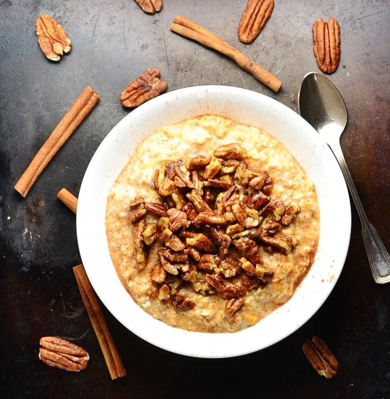 Top down view of sweet potato overnight oats with maple pecans on top of dark surface with spoon and cinnamon sticks and pecans scattered about.