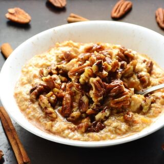 Side view of sweet potato overnight oats with pecans and spoon in white bowl on top of dark table with cinnamon sticks and pecans scattered about.