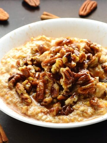 Side view of sweet potato overnight oats with pecans and spoon in white bowl on top of dark table with cinnamon sticks and pecans scattered about.