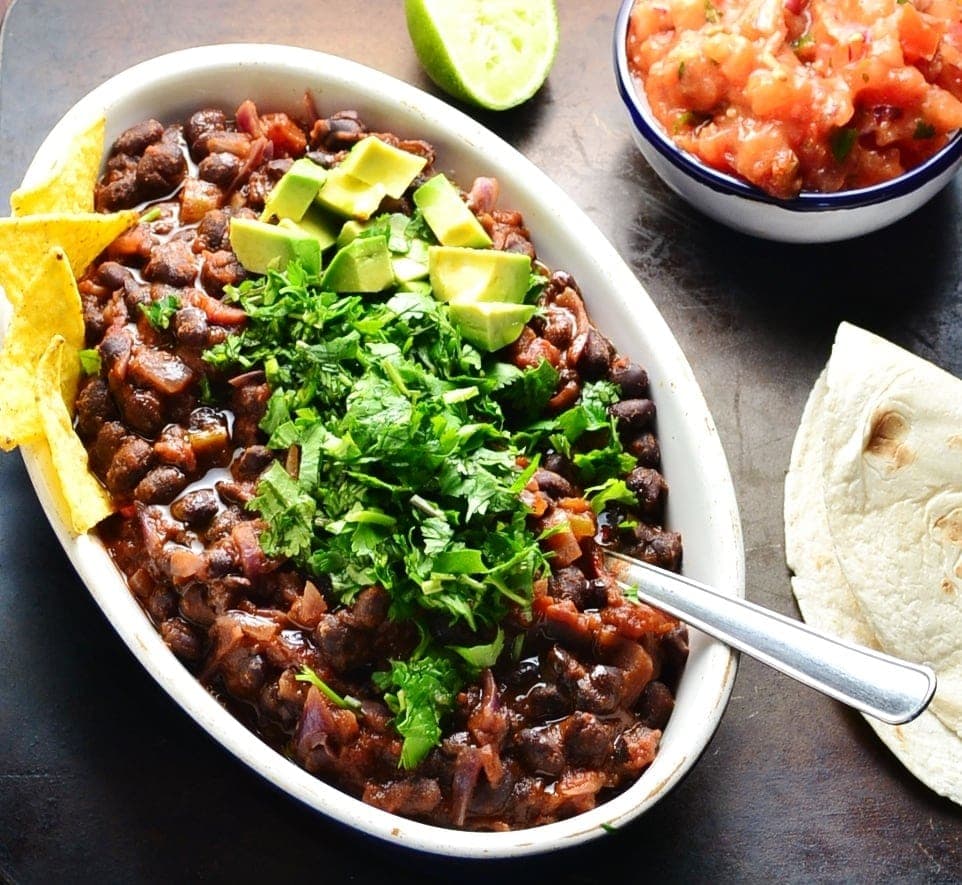 Close-up view of black bean stew in white oval dish garnished with chopped cilantro, avocado and nachos, with lime, salsa in blue dish and tortilla on dark brown surface.