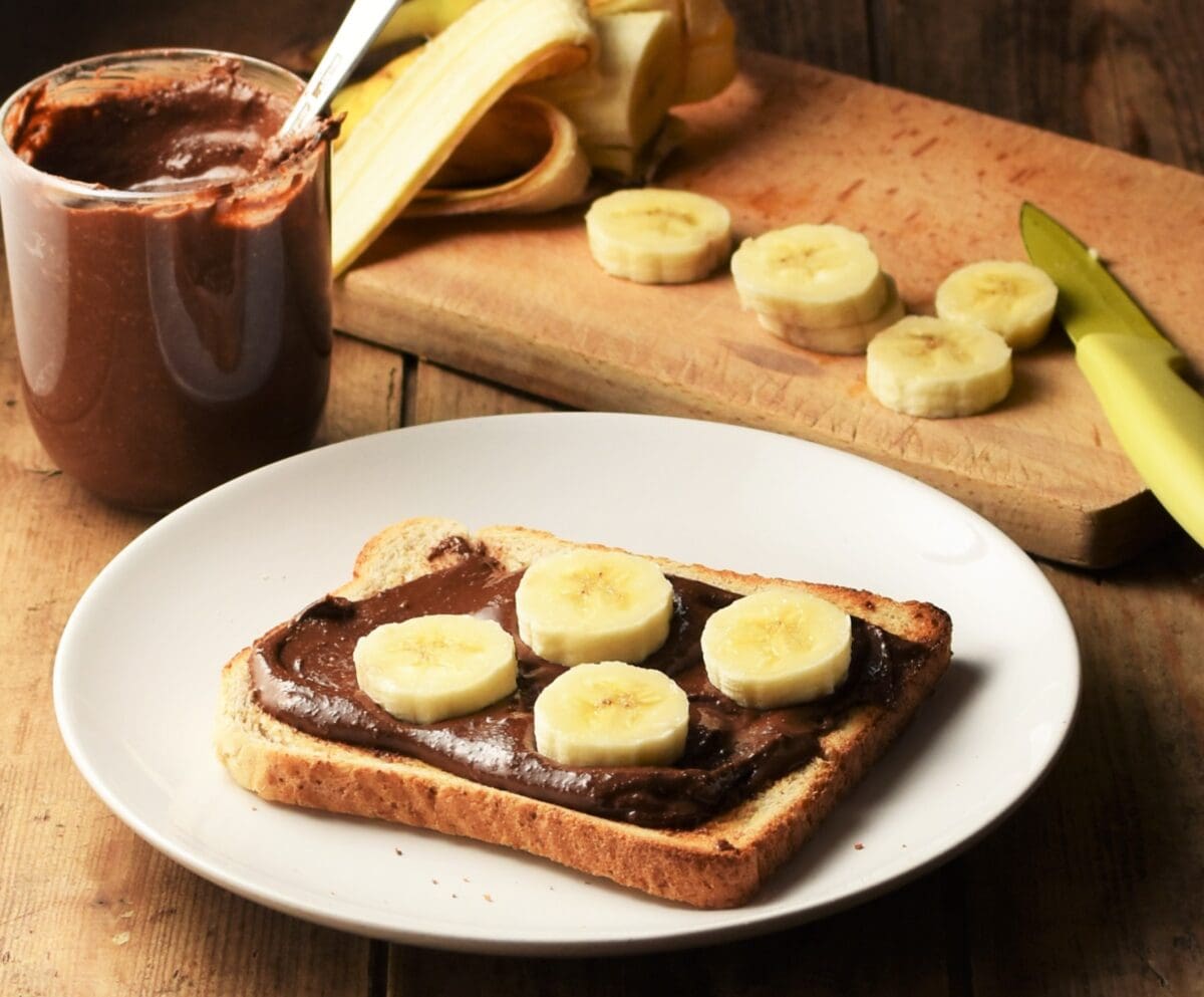Side view of chocolate spread on toast with banana on white plate, spread in cup and chopped banana in background.