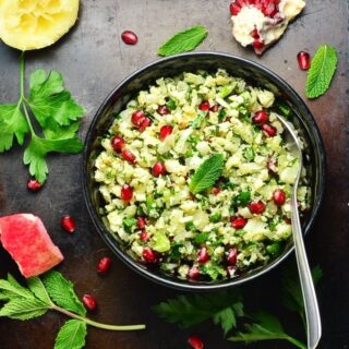Top down view of roasted cauliflower tabbouleh salad with pomegranate seeds and spoon in black bowl, with herbs and pomegranate seeds scattered around.