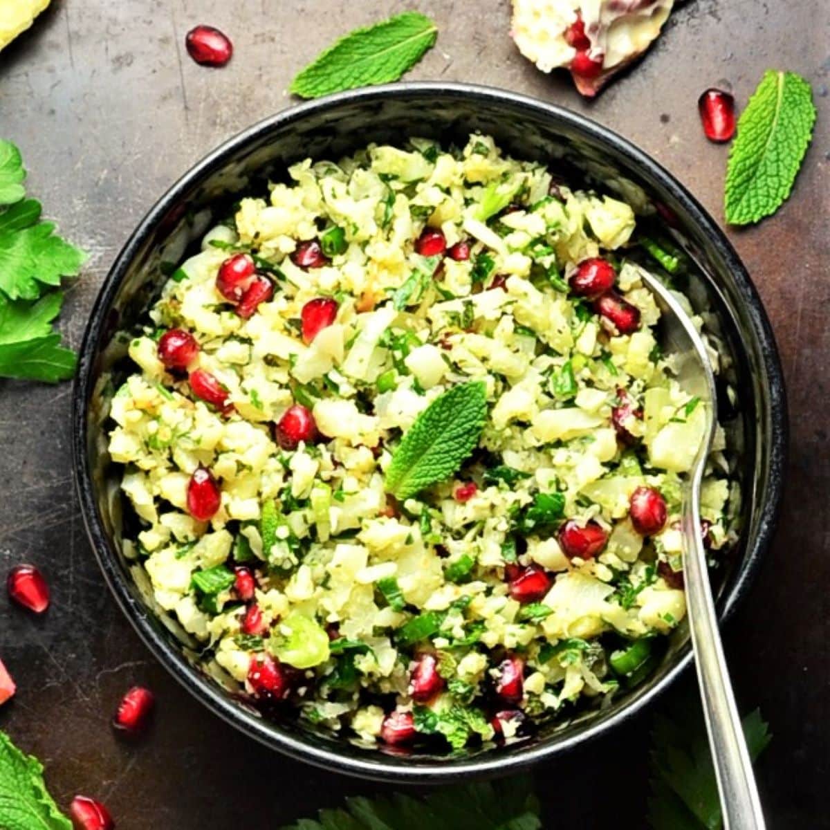 Cauliflower tabbouleh salad with herbs and pomegranate in black bowl with spoon.