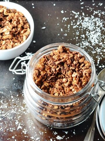 Top down view of chocolate granola in open jar with spoon to right and sea salt on top of dark shiny surface.
