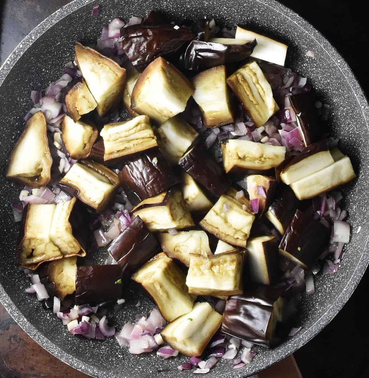 Pieces of roasted eggplant with chopped red onion in pan.