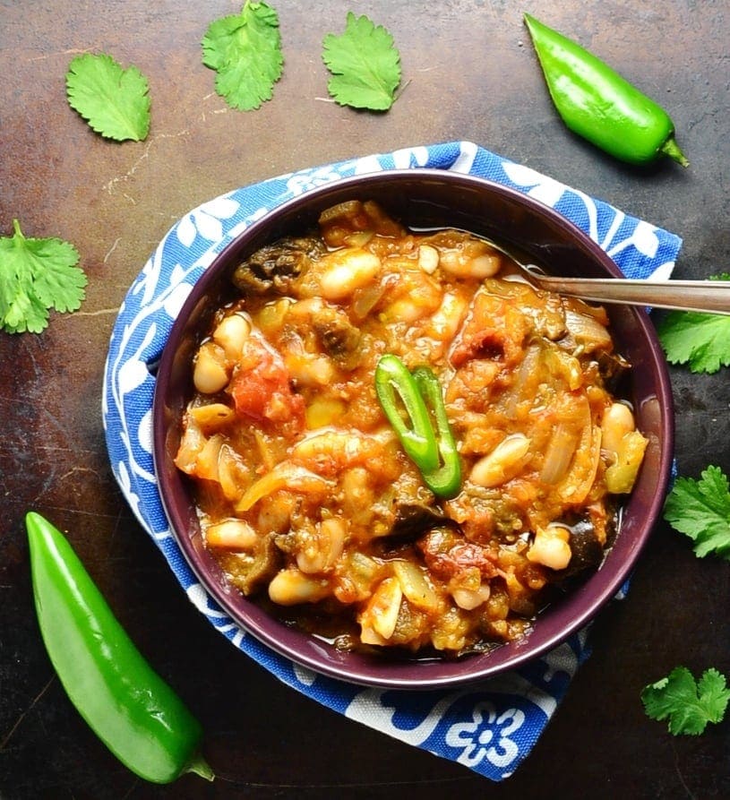 Top down view of eggplant in tomato sauce in purple bowl with spoon wrapped in blue-and-white cloth with green chillies and cilantro leaves on dark brown surface.