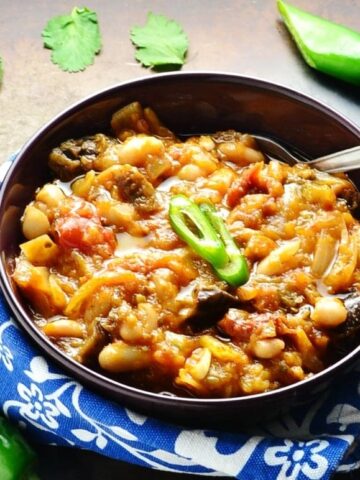 Eggplant casserole with spoon in purple bowl wrapped in blue-and-white cloth with green chillies and cilantro leaves on dark brown surface.