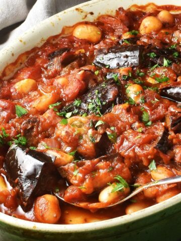Side view of eggplant casserole with beans and rich tomato sauce in oval dish with spoon.