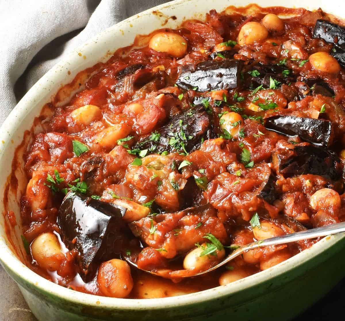 Side view of eggplant casserole with beans and rich tomato sauce in oval dish with spoon.