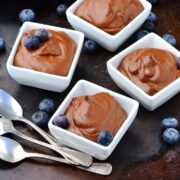 Chocolate pudding in 4 white square dishes, with 3 spoons and blueberries on top of dark surface.