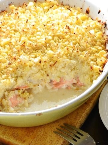 Healthy fish pie with cauliflower topping in white oval casserole dish on wooden board with fork and partial view of white plate with pie.