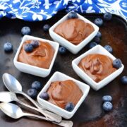 Side view of chocolate pudding in 4 white square dishes topped with blueberries on top of dark surface with 3 spoons, blueberries and blue-and-white cloth at top.