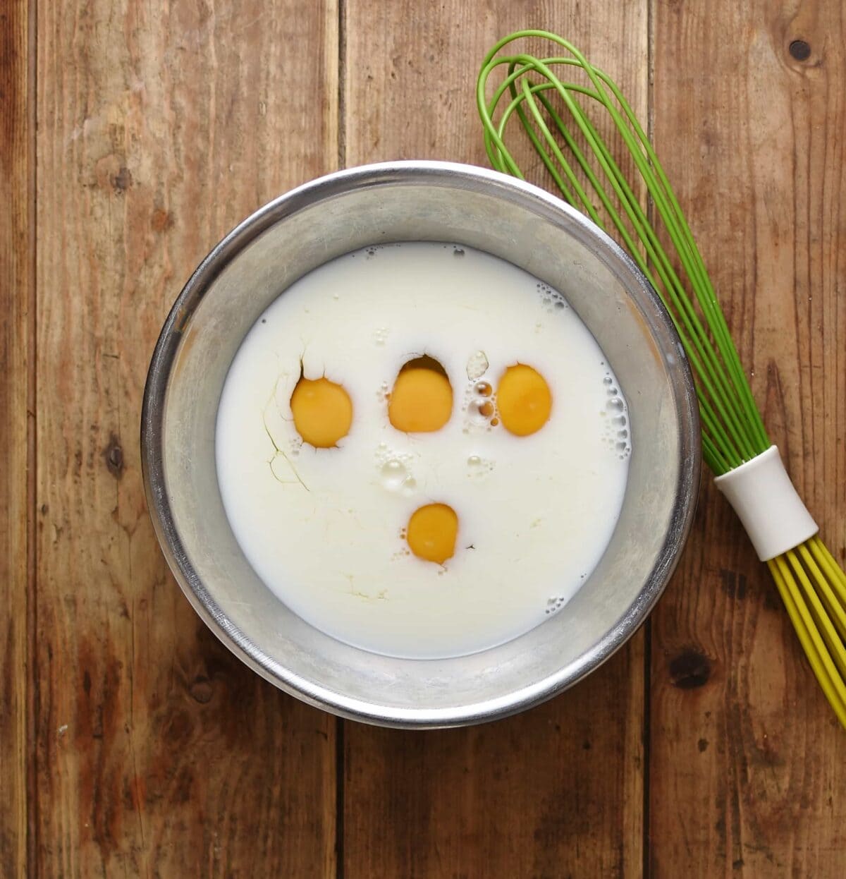 4 eggs with milk inside metal bowl with green whisk to the right.