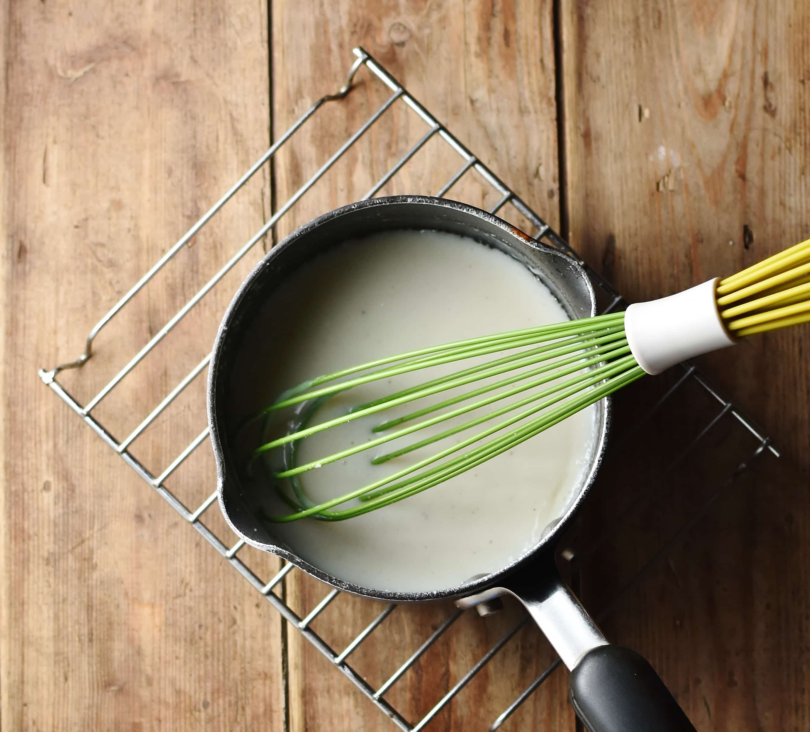 White sauce in small saucepan with green whisk on top of cooling rack.