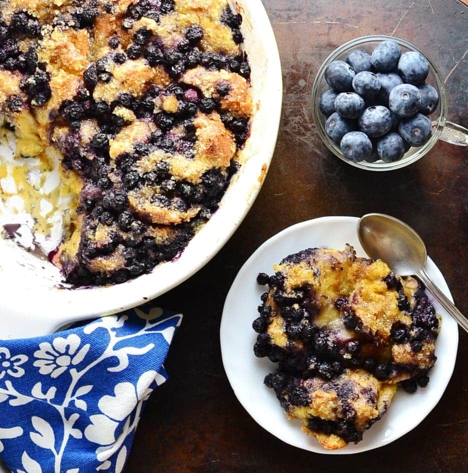 Top down view of blueberry french toast casserole on white plate with spoon, casserole in white oval dish, cup with fresh blueberries and blue-and-white cloth.