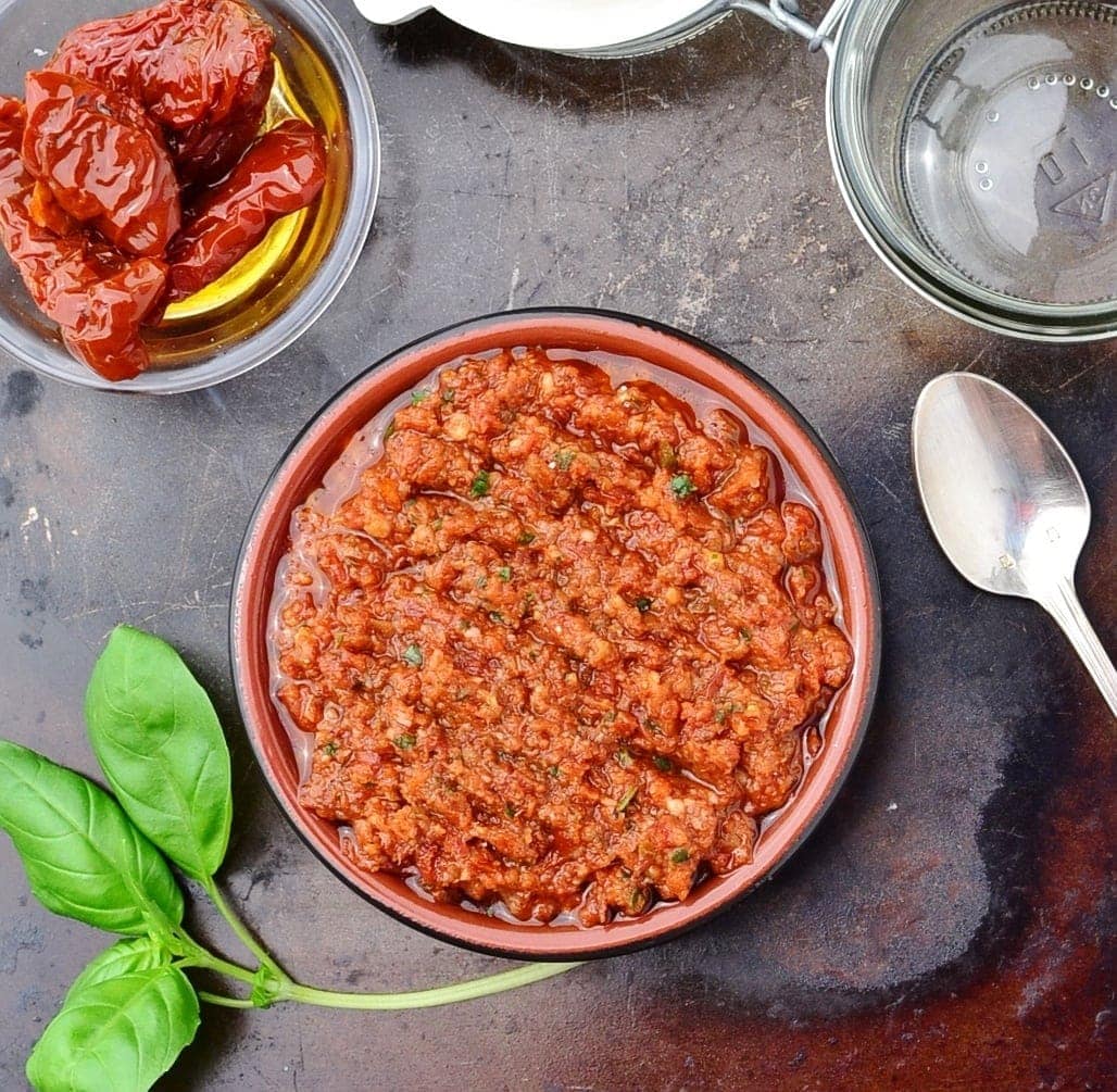Top down view of sun dried tomato pesto sauce in round dish on top of dark surface with basil leaves, spoon, open jar and sun dried tomatoes in small dish.