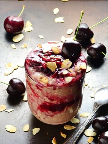 Cherry overnight oats with fresh cherries, almond flakes and spoon on dark brown surface.