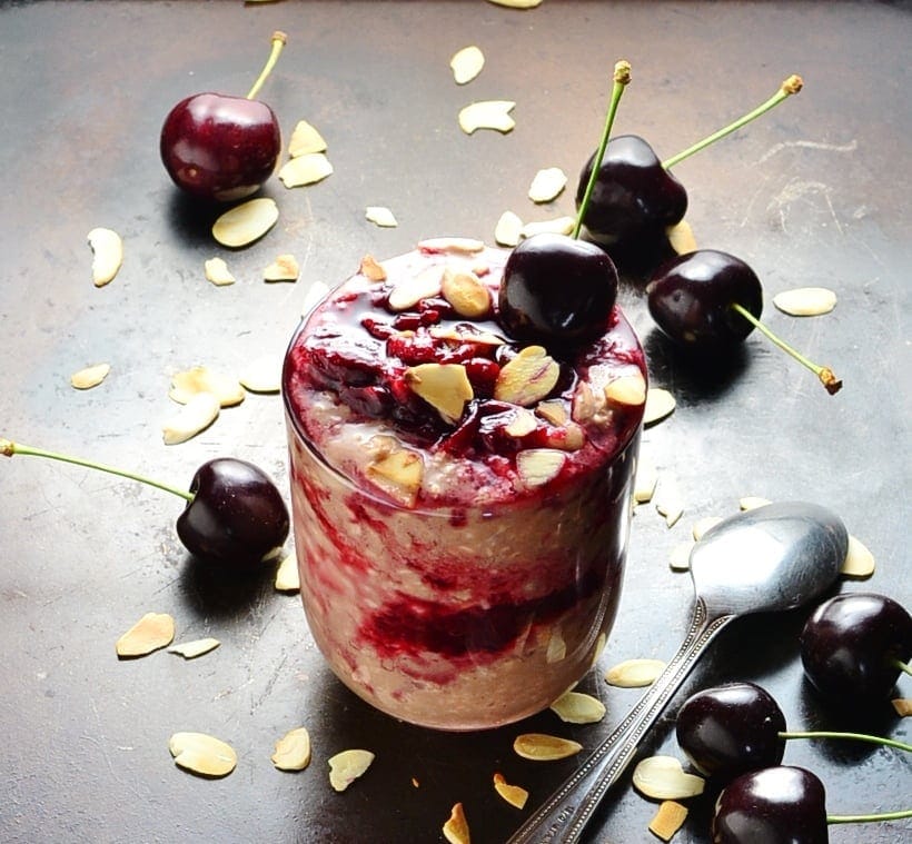 Cherry overnight oats with fresh cherries, almond flakes and spoon on dark brown surface.