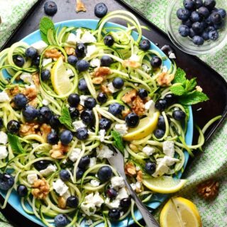Top down view of blueberry feta salad with spiralized zucchini and lemon wedges on top of blue plate on dark tray, with blueberries in small dish and green cloth in background.
