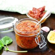 Top down view of sun dried tomato pesto in round dish on top of dark surface with basil leaves, spoon, open jar and sun dried tomatoes in small dish.