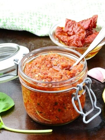 Top down view of sun dried tomato pesto in round dish on top of dark surface with basil leaves, spoon, open jar and sun dried tomatoes in small dish.