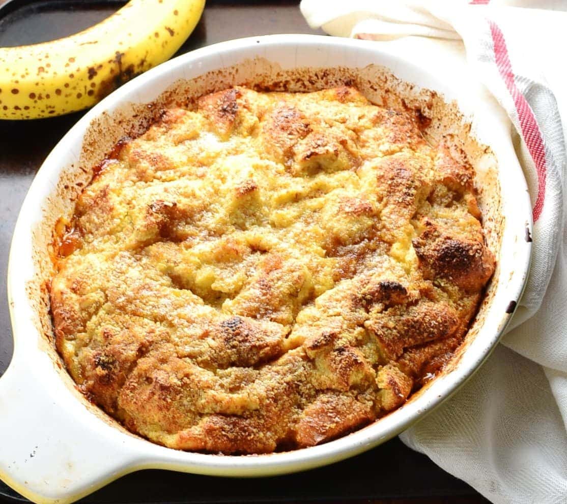 Banana french toast bake in white oval dish with ripe banana at top and white cloth with red stripe to right.