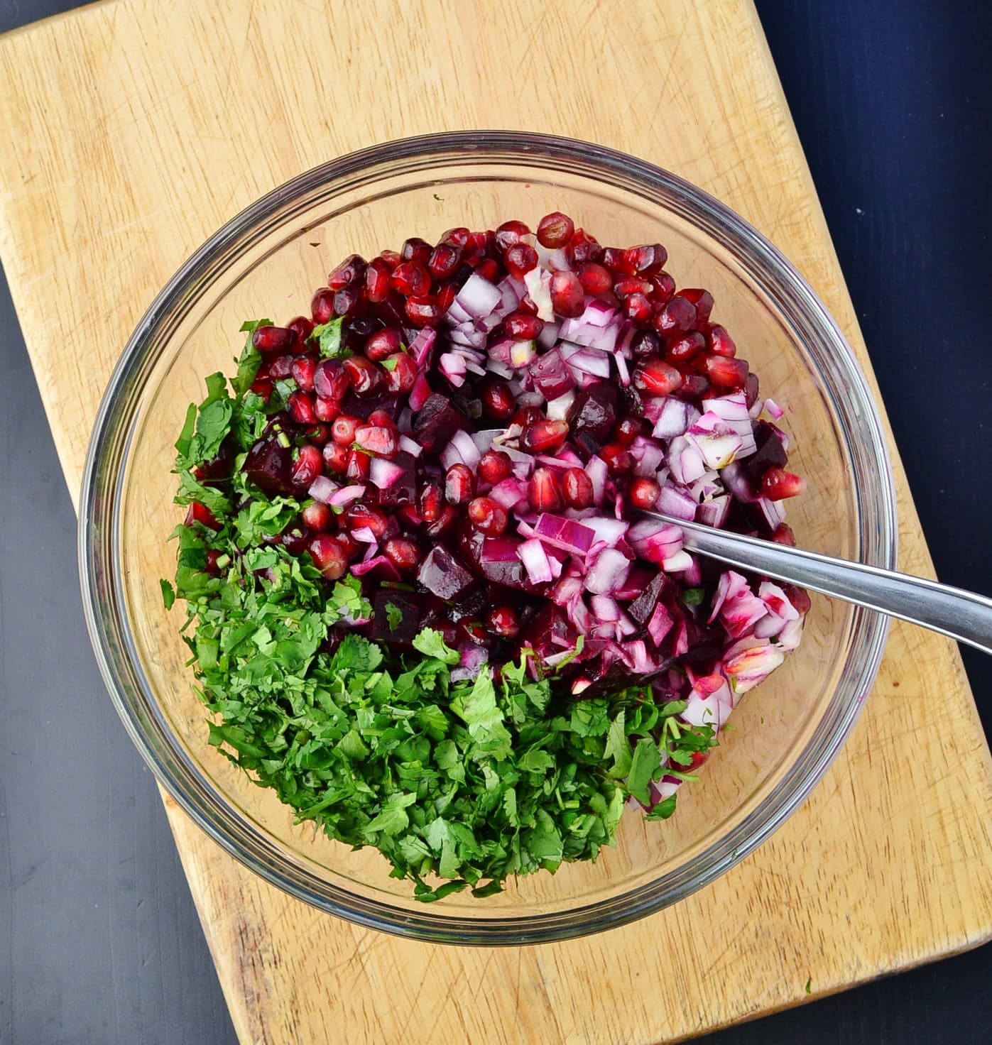 Chopped onions, pomegranate seeds and cilantro in see through bowl with spoon on wooden board.