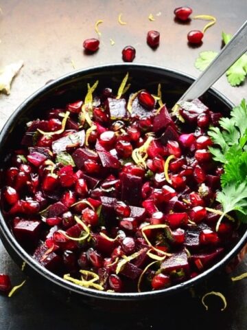 Beetroot salsa with pomegranate in black bowl with spoon and lemon, cilantro and pomegranate seeds scattered on dark brown surface.