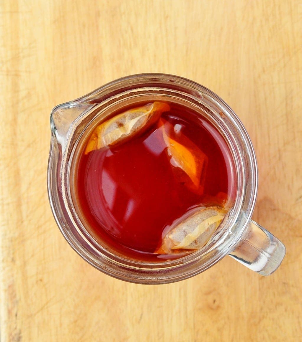 Top down view of red rooibos iced tea with tea bags in see-through jug on top of wooden table.
