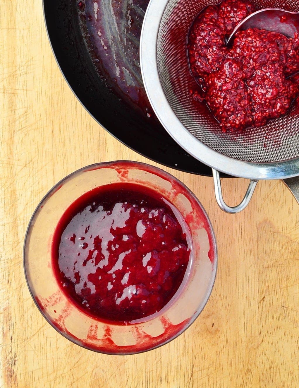 Top down view of raspberry sauce in small bowl and raspberry pulp with spoon inside sieve.