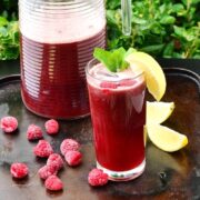 Side view of rooibos iced tea with lemon wedge and mint leaves in tall glass, with frozen raspberries and lemon wedges on top of brown tray, and jug of iced tea in background.