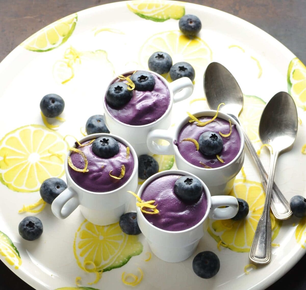 Blueberry cream cheese dessert in 4 white cups with garnish of blueberries and lemon zest, with 2 spoons on white plate with lemon pattern.