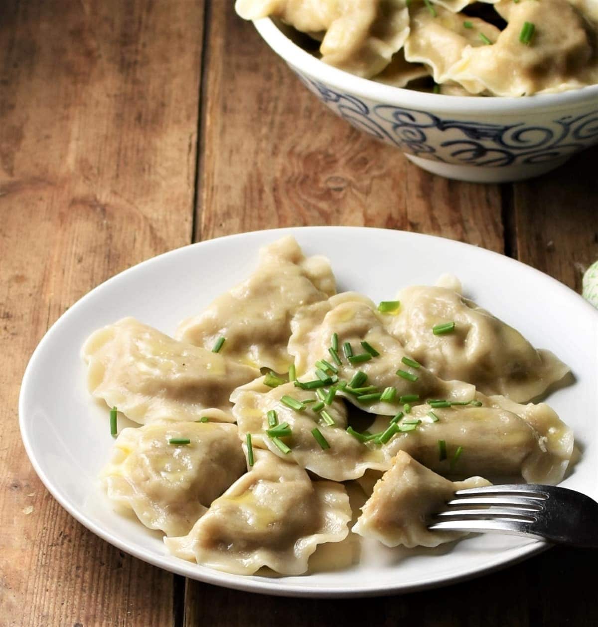 Side view of pierogi on top of plate with pierogi in bowl in background.