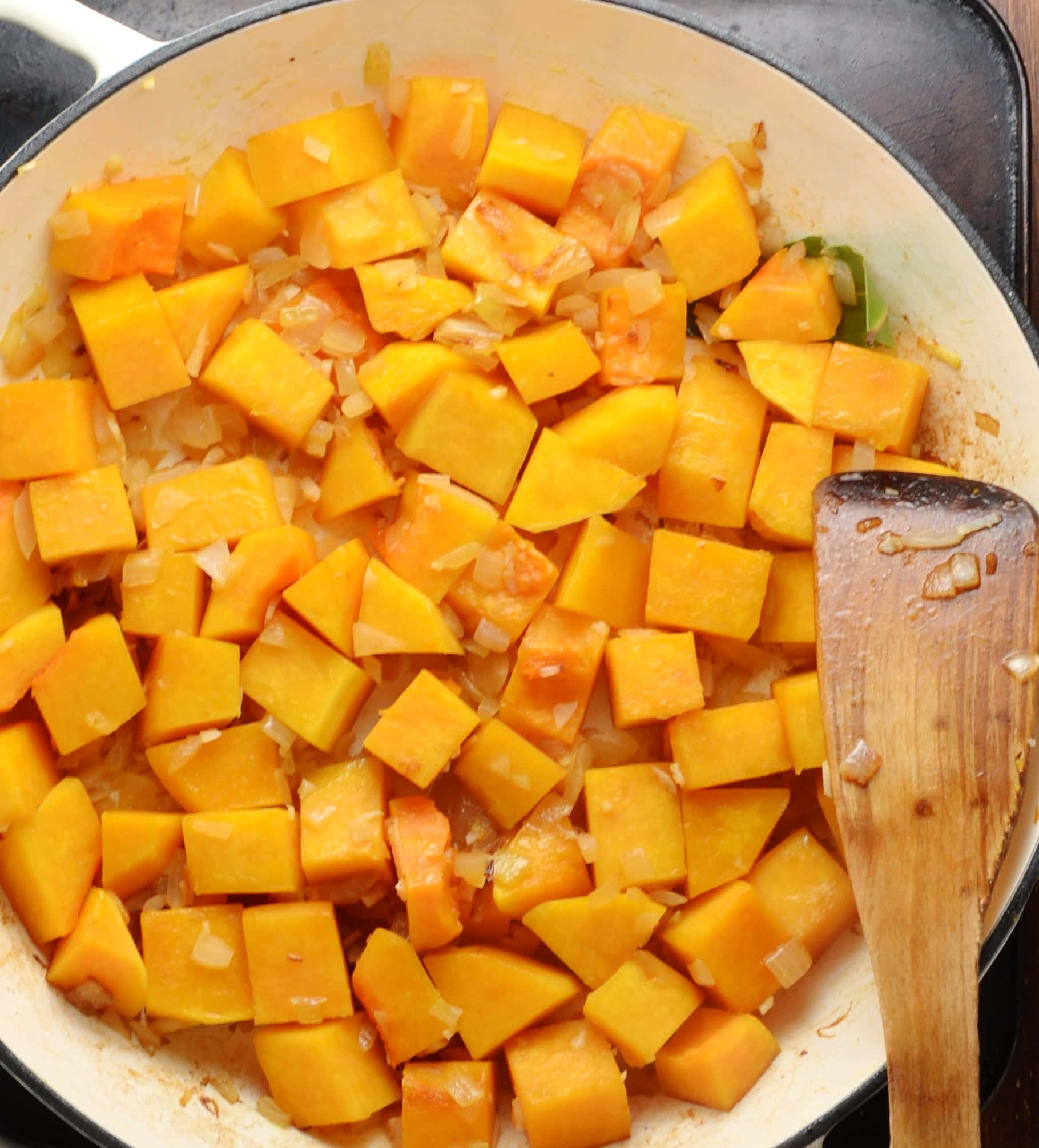 Top down view of cubed butternut squash with onions in large shallow white dish with wooden spoon.