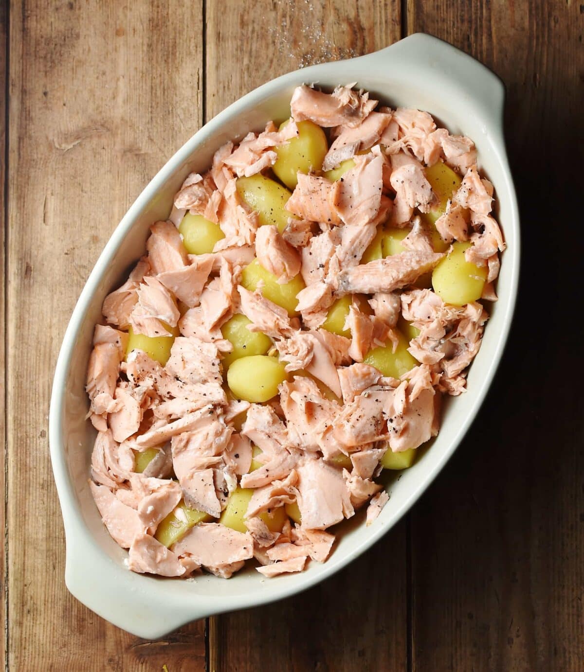 Shredded cooked salmon on top of potatoes in oval, white casserole dish.