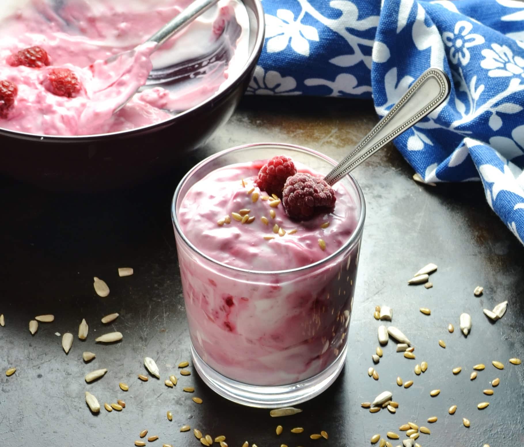 Flavoured raspberry yogurt in glass with spoon, bowl and blue-and-white cloth in background.