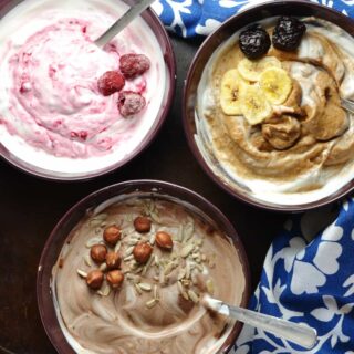 Flavoured mocha, raspberry, prune yogurt in purple bowls with spoons and blue-and-white clothe on dark surface.
